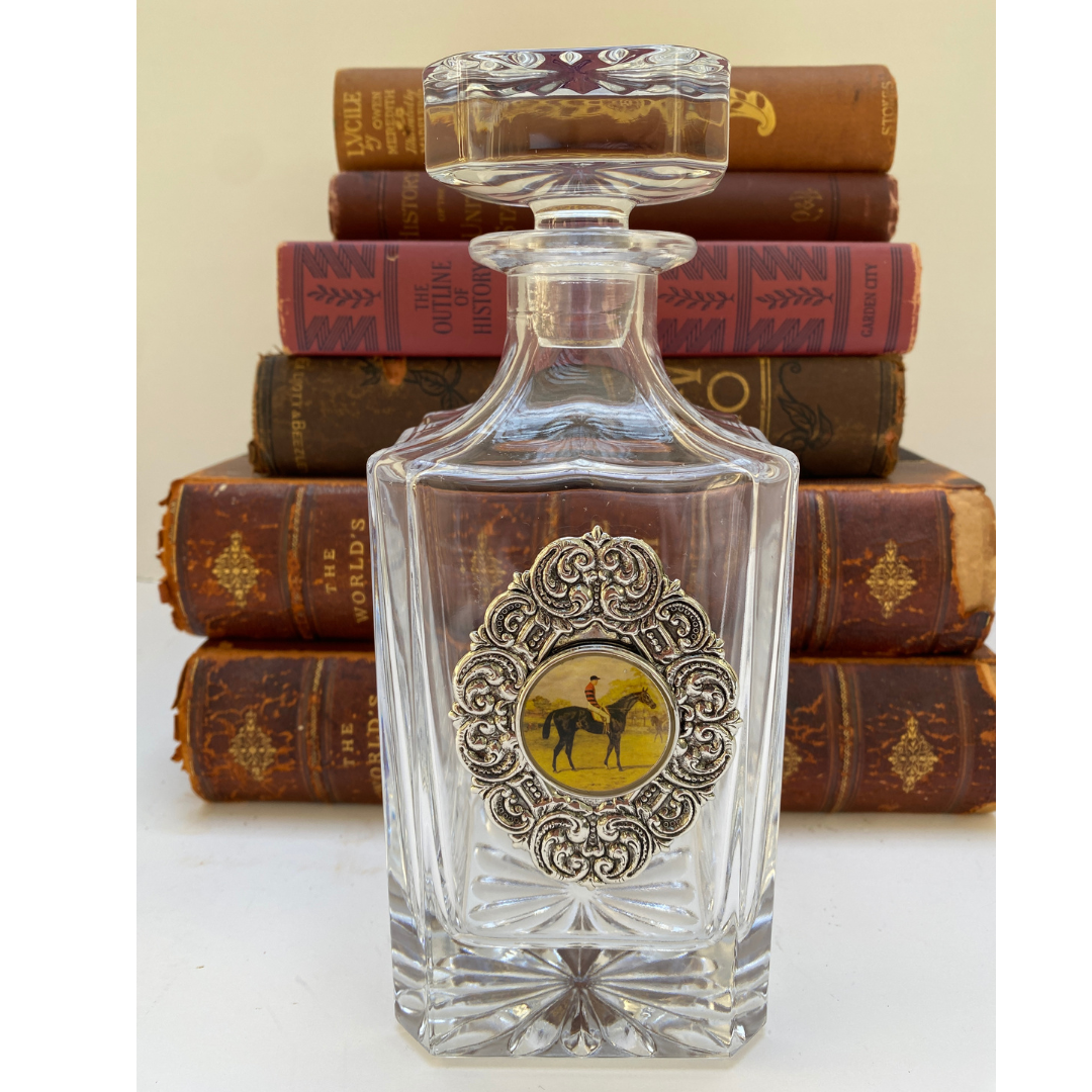 A clear glass decanter with a silver medallion featuring a vintage racehorse design, placed on a surface with books in the background.