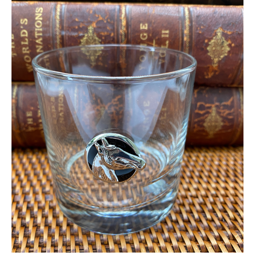 A clear whiskey bourbon glass with a silver horse-head medallion on the side, held by a person.