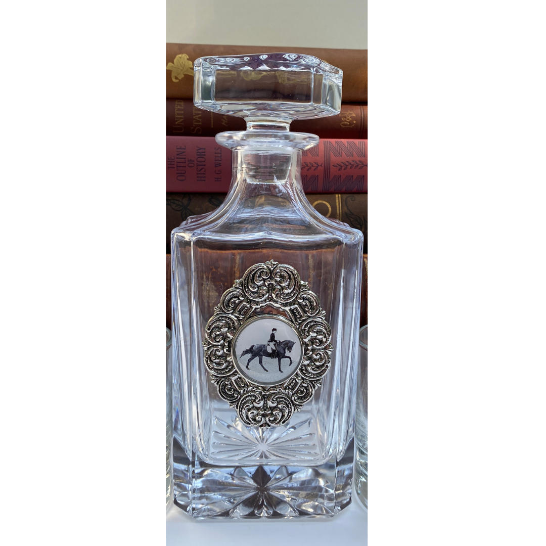 A clear glass decanter with a silver medallion embellishment featuring a dressage rider in the center, placed on a surface with books in the background.
