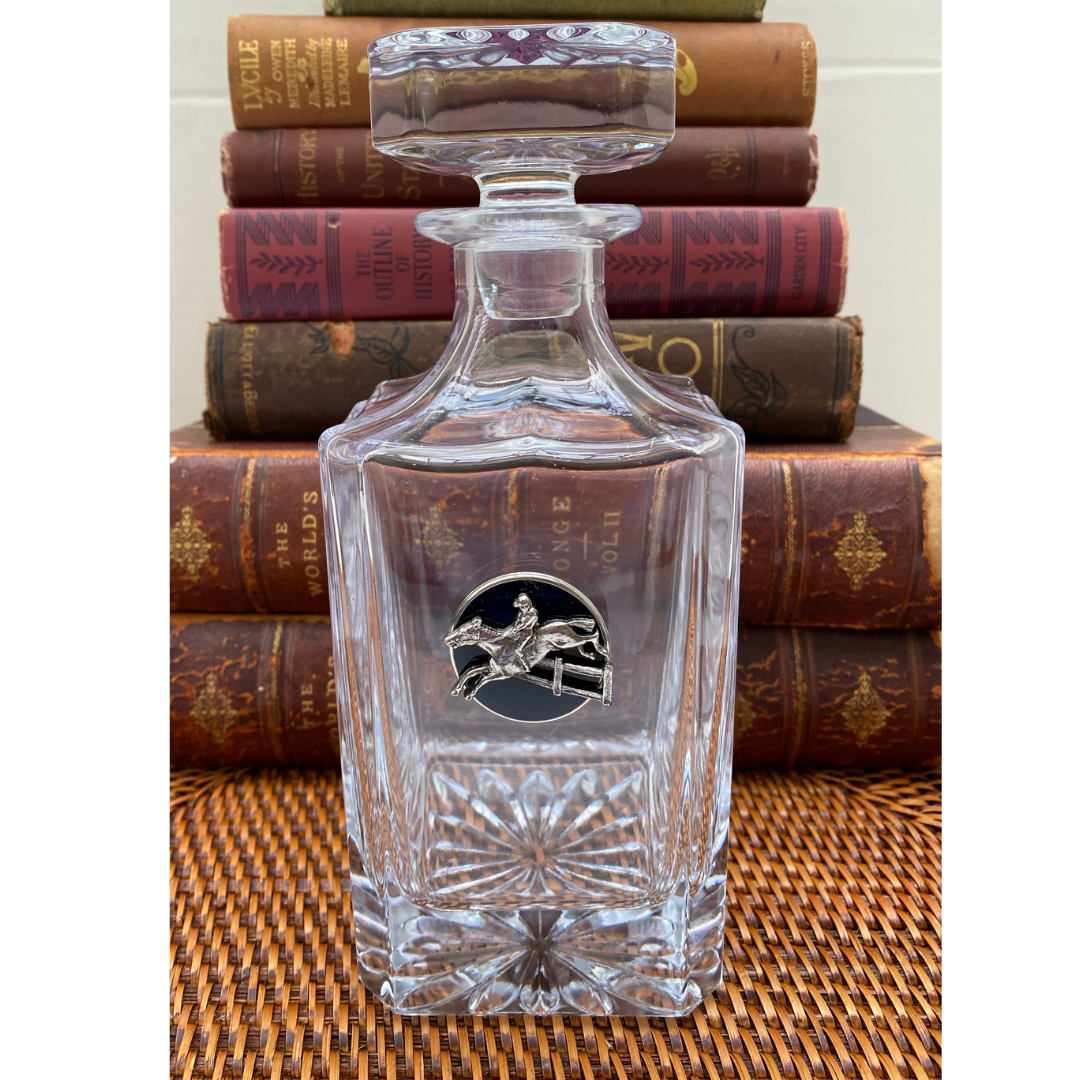 A clear square-shaped decanter with a round medallion featuring a silver hunter jumper design, displayed on a wicker surface against a backdrop of stacked vintage books.