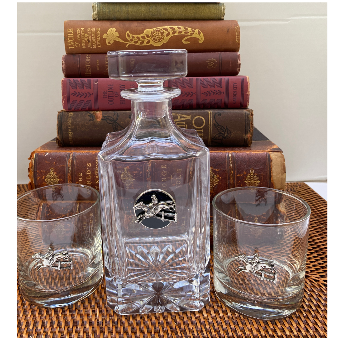 A clear glass decanter with a round medallion featuring a black enamel and silver hunter jumper design, displayed in front of a backdrop of books and beside two glasses with a woven tablecloth.