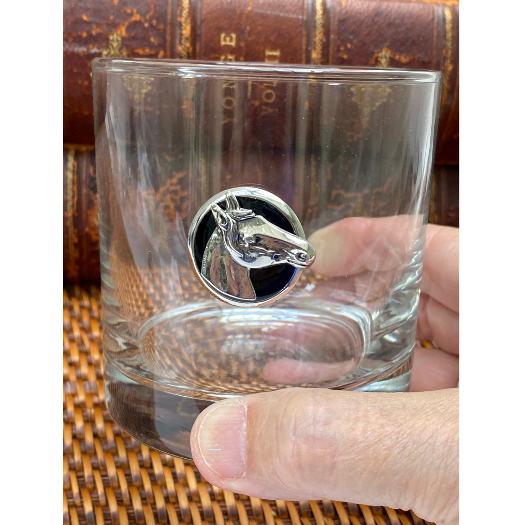 A clear whiskey bourbon glass with a silver horse-head medallion on the side, held by a person.