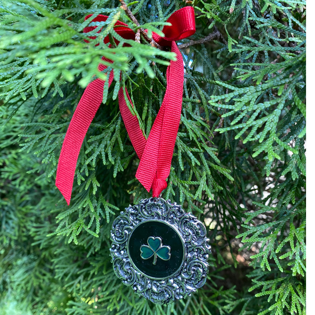 Decorative ornament with a shamrock on a red ribbon hanging on a green tree.
