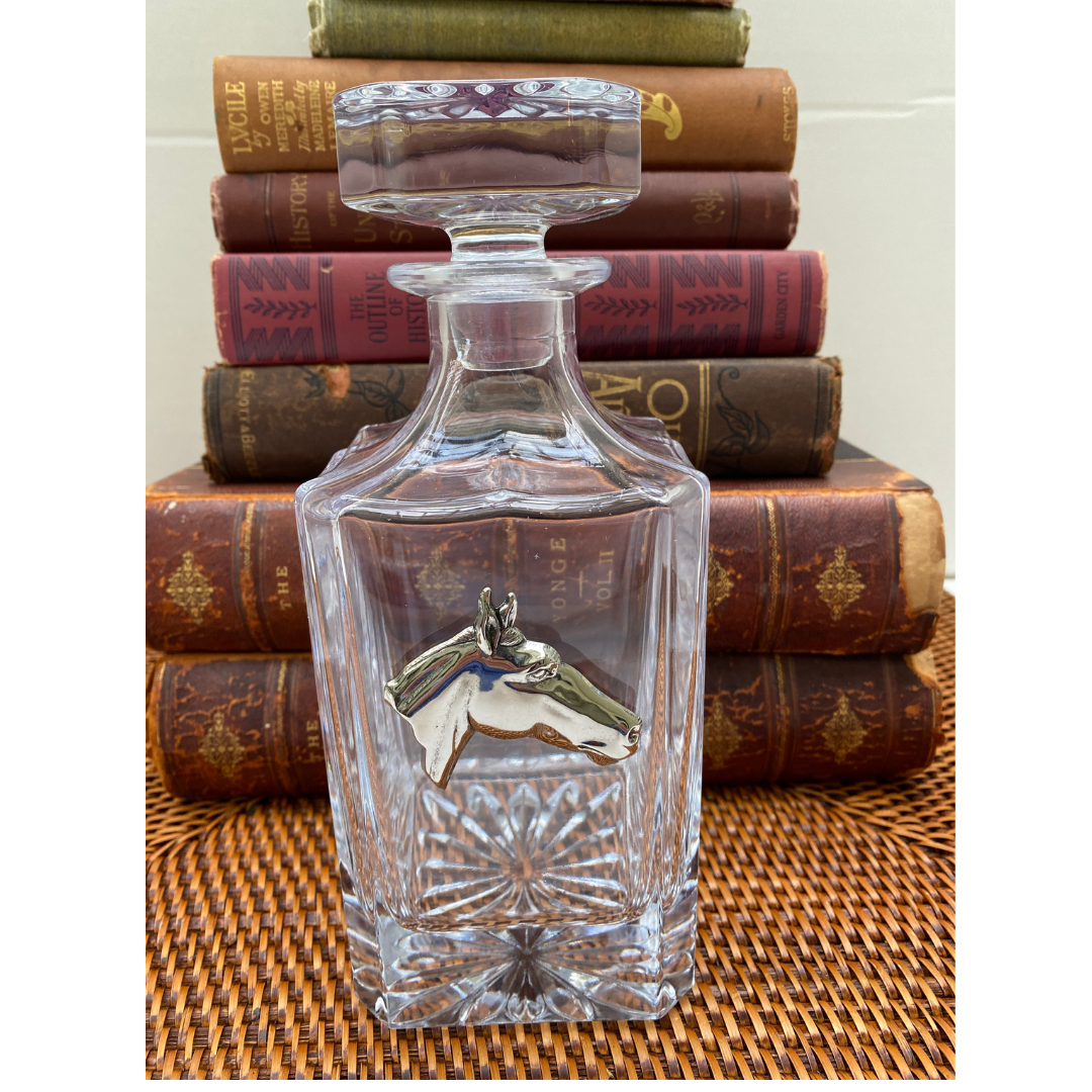 A clear glass decanter with a silver medallion of a horse head, placed on a surface with books in the background.