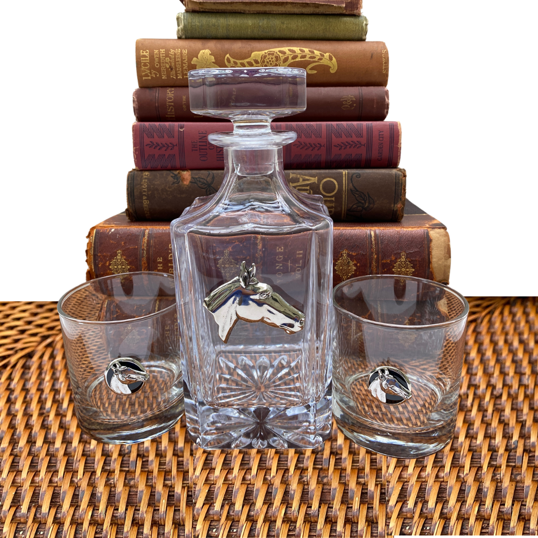 A clear glass decanter with a silver medallion of a horse head, placed on a surface with books in the background.