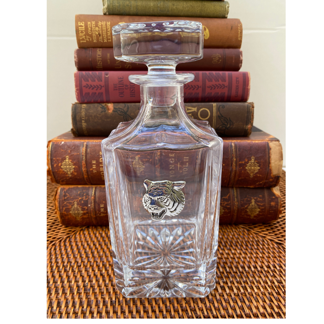 A clear glass decanter with a silver tiger head medallion, placed on a stack of books.