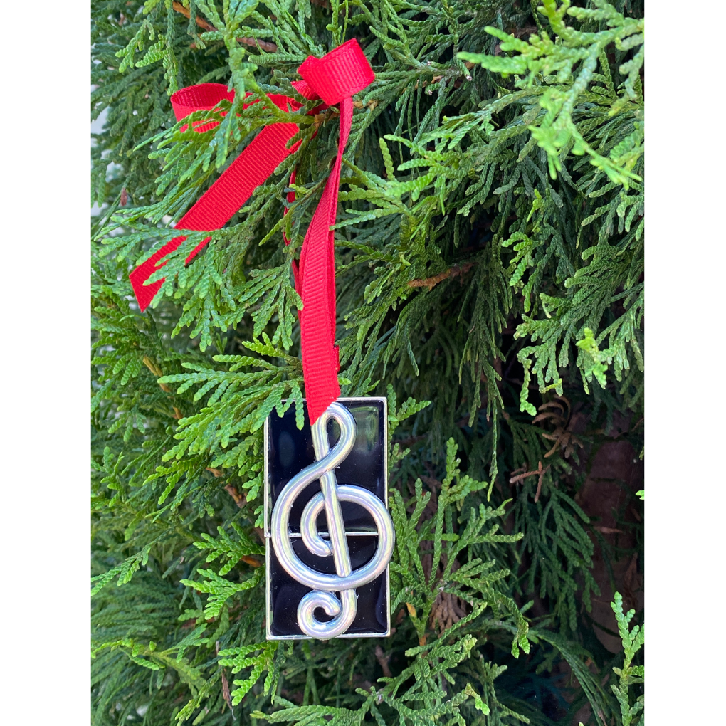 Decorative ornament with a treble clef symbol on a red ribbon against a green Christmas tree background.