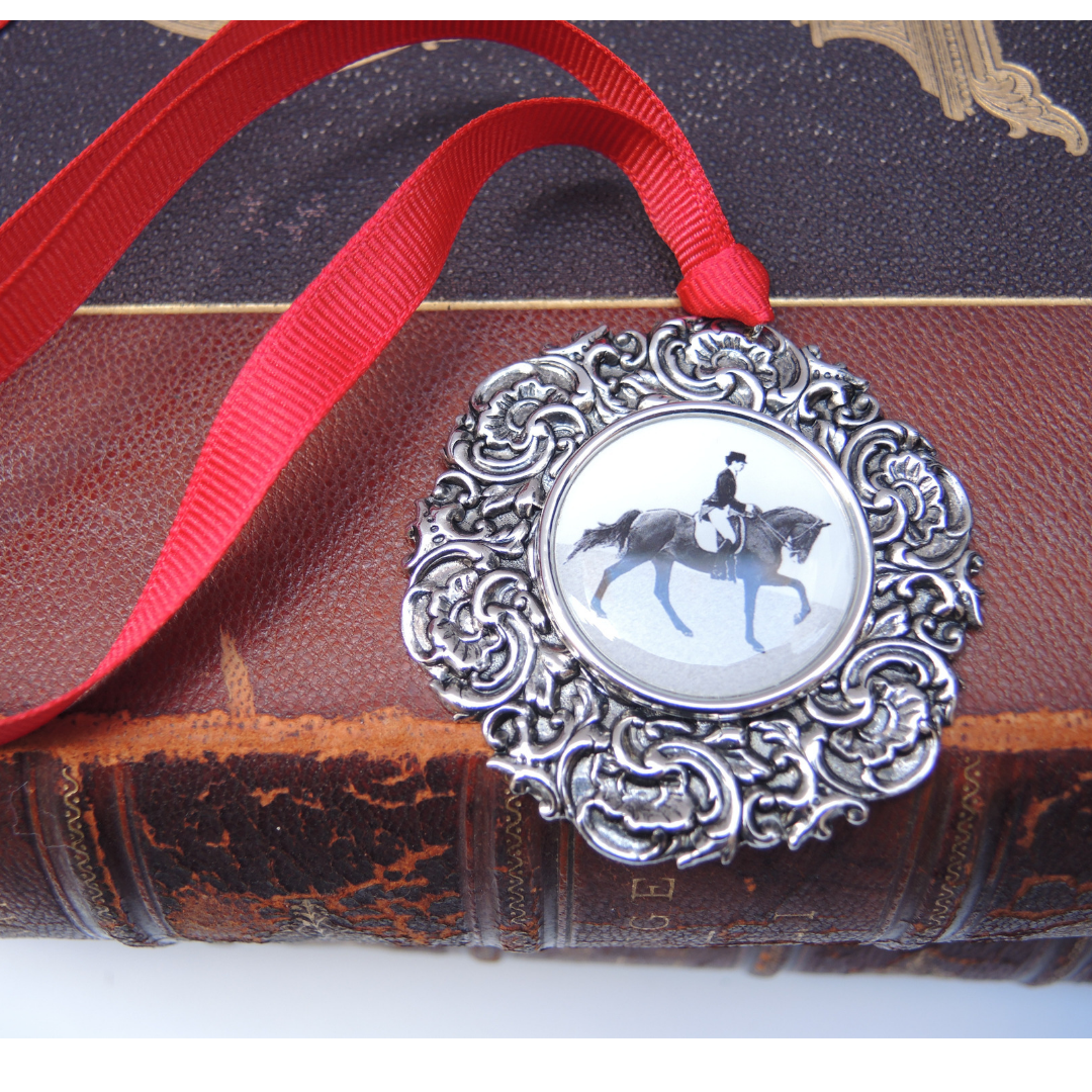 Dressage Christmas ornament featuring the rider and horse in the center of a vintage-inspired silver medallion. It is hung with a red grosgrain ribbon
