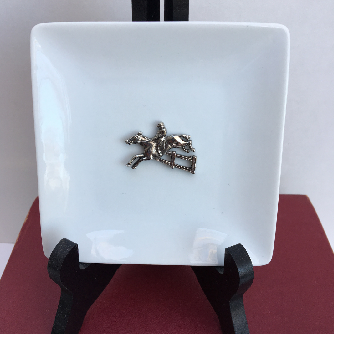 A white porcelain trinket tray with a silver horse jumper medallion in the center, displayed on a black stand.
