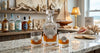 A clear glass decanter and two matching rocks glasses with a scallop shell motif, filled with amber liquor, displayed on a marble countertop in a nautical-themed bar setting.