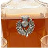 A close-up of a clear glass decanter filled with amber liquid, featuring a silver-toned metal thistle and laurel wreath emblem on the front.