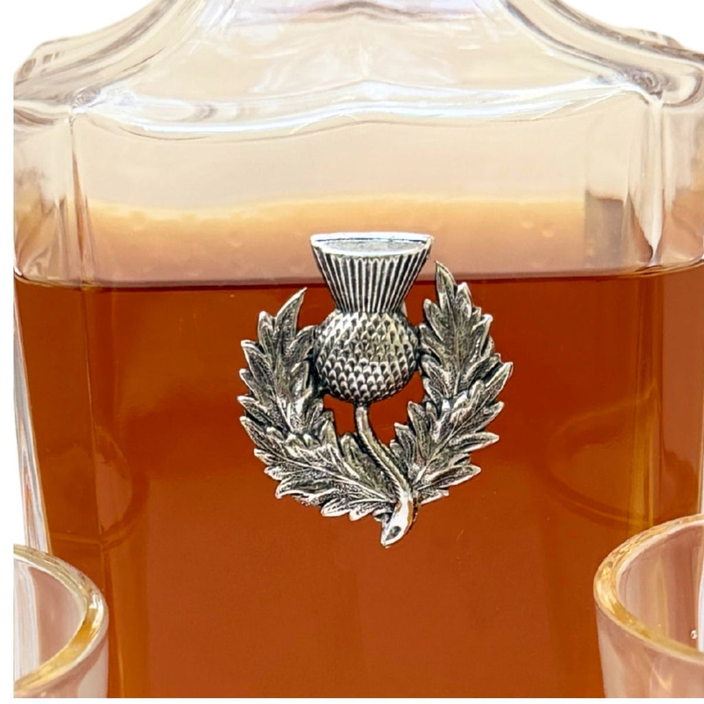 A close-up of a clear glass decanter filled with amber liquid, featuring a silver-toned metal thistle and laurel wreath emblem on the front.