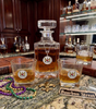 A custom clear glass decanter and two matching rocks glasses with a circular Saint Louis Bourbon Street Cemetery emblem, filled with amber liquor, displayed on a granite bar counter with Mardi Gras beads, a fleur-de-lis, and a sign reading Bourbon Street.