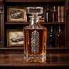 A clear glass whiskey decanter with a stopper, filled with amber liquid, featuring an ornate silver metal emblem on the front. The decanter is set on a dark wooden table with a matching wooden shelf in the background containing framed vintage car photos and other bottles.