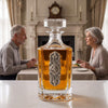 A clear glass decanter filled with amber liquor and decorated with a silver ornate emblem, placed on a white table in a dining room with an older couple seated in the background.