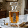 A clear glass decanter filled with amber liquor, featuring an ornate silver-toned metal emblem on the front, set on a marble countertop with a small glass, jigger, and vintage book in a warm indoor setting.
