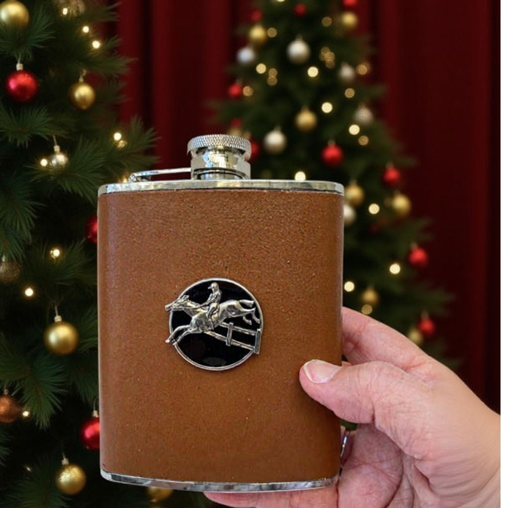 Brown leather flask with silver emblem held in front of a decorated Christmas tree.  Gift for steeplechase rider and hunter jumper  fan.