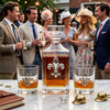 A clear glass decanter and two matching rocks glasses with silver fleur-de-lis emblems, filled with amber liquor, displayed on a marble table in front of a group of elegantly dressed people at an outdoor social event.