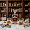 A luxury decanter and two matching coupe glasses with silver fleur-de-lis accents, filled with amber liquor, arranged on a white marble table in front of a dark wood bookshelf.