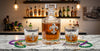 A fleur de lis clear glass decanter and two matching rocks glasses with fleur-de-lis engravings, filled with amber liquor, arranged on a marble countertop with Mardi Gras beads and a blurred bar shelf background.
