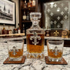 A clear fleur de lis glass whiskey decanter and two matching lowball glasses with fleur-de-lis cutouts, filled with amber whiskey, arranged on a marble countertop in a bar setting.
