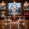 A clear fleur de lis  glass decanter and two matching lowball glasses with silver fleur-de-lis emblems, filled with amber liquor, are arranged on a wooden table in a bar-like interior with framed pictures and a city skyline window behind them.