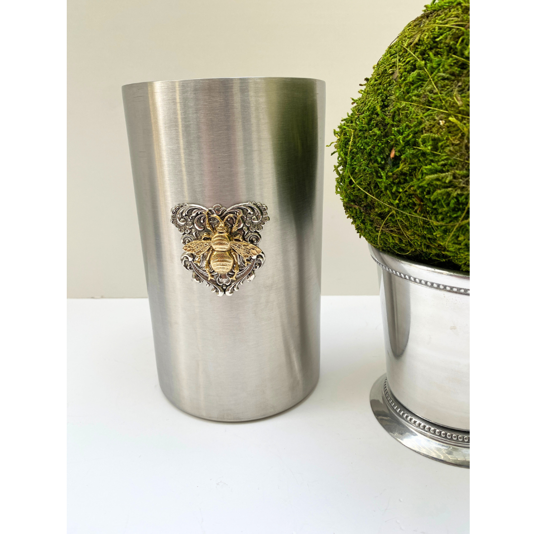A stainless steel wine cooler with a silver bee medallion design, displayed next to a green artificial plant.