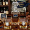 A clear glass Happy Birwhiskey decanter with a stopper and two matching rocks glasses, all filled with amber liquor, arranged on a wooden bar counter. The glasses have oval silver-and-white decorative medallions, and the decanter has a small blue label reading Happy Birthday