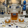 A clear glass  Happy Birthday whiskey decanter and two matching lowball glasses filled with amber liquor, arranged on a marble table with confetti and napkins. The scene is decorated for a birthday with a blue and gold 'HAPPY BIRTHDAY' banner, gift boxes, bottles on shelves, and blue and silver balloons in the background.