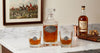 A horse and hound whiskey decanter and two matching rocks glasses with silver medallion engravings, displayed on a white marble countertop next to a bottle of Kentucky Reserve whiskey and a copper jigger.