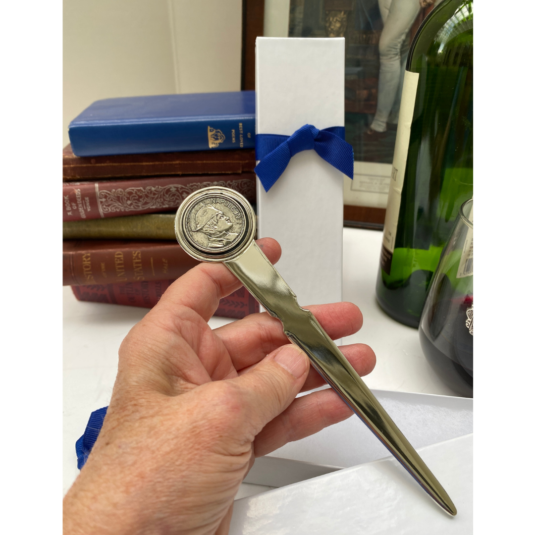 A silver-colored letter opener featuring a silver Napoleon coin at the top, held in a person's hand, with a white box and blue ribbon in the background.