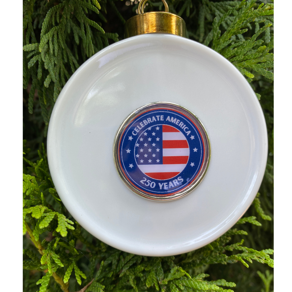 White ornament with a circular design featuring an American flag and text, set against a green leafy background.