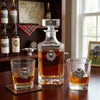 A clear glass decanter and two matching rocks glasses with silver thistle emblems, filled with amber liquor, arranged on a wooden table in a warm indoor setting with bottles and framed decor in the background.