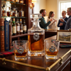 A clear glass whiskey decanter and two matching rocks glasses with silver thistle and wreath engravings, filled with amber whiskey, displayed on a wooden table in a warmly lit bar or lounge setting with blurred people and bottles in the background.