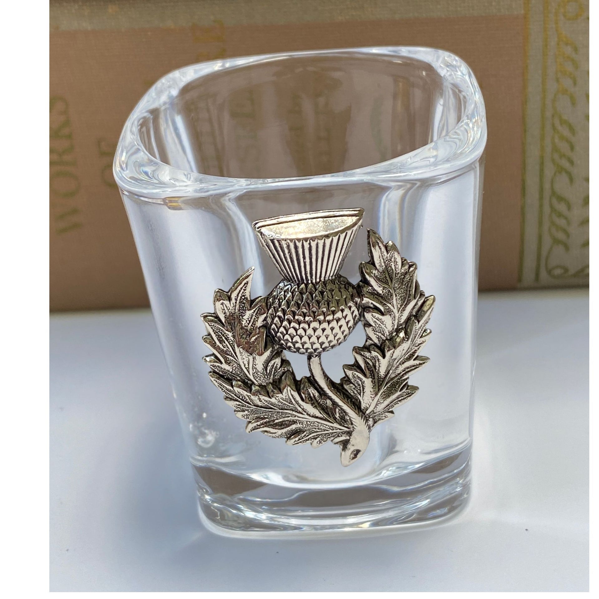 Clear glass shot glass with a metallic thistle emblem on a white surface.