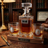 A clear glass  Thank You theme whiskey decanter and two matching rocks glasses on a wooden presentation tray, filled with amber liquor. The set is displayed in a warm, rustic library setting with bookshelves, a brass lamp, and a pen on a leather pad.