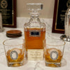 A Thank you decanter set  clear glass decanter filled with amber liquid stands on a marble surface, flanked by two matching clear glasses with large ice spheres and a small card in front. The decanter has an ornate silver-toned label reading THANK YOU, and the glasses have matching decorative circular cutouts.