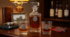 A clear glass whisky decanter and two matching rocks glasses with engraved silver thistle emblems, filled with amber whisky, displayed on a wooden table with a lamp, books, and whisky bottles in the background.