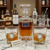 A Welcome Decanter Set  clear glass decanter and two matching rocks glasses with oval silver-and-white medallion engravings, styled on a marble countertop with whiskey bottles, a shaker, and bar tools in the background. A small sign reads 'Welcome to the Neighborhood!'.