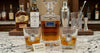 A Welcome Decanter Set whiskey decanter and two rocks glasses filled with amber liquor on a marble countertop, with liquor bottles and bar tools blurred in the background. A small sign in front reads 'Welcome to the Neighborhood!'.