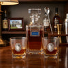 A Welcome Decanter Set  clear glass decanter and two matching lowball glasses with ornate silver oval medallions, filled with amber liquor, arranged on a wooden bar counter with bottles and bar accessories blurred in the background.