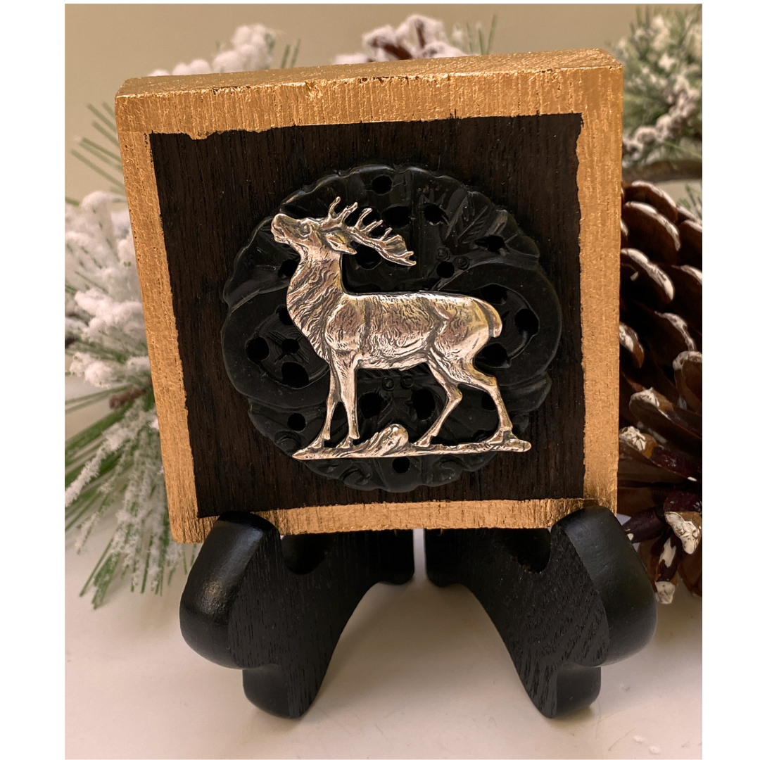 A wooden art piece featuring a silver stag deer medallion in the center, with dark carved jade edges and a gold leaf painted back, displayed on a black stand.