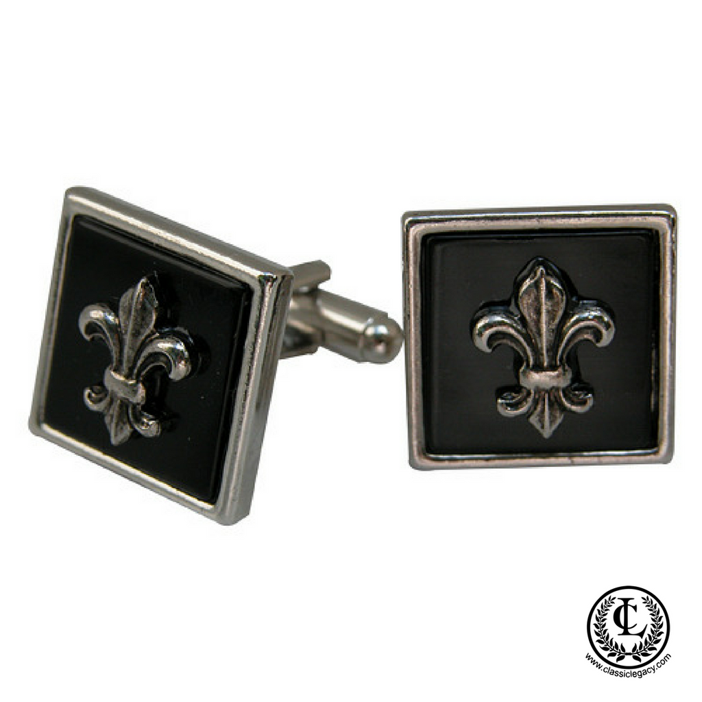 A pair of silver cuff links with a black enamel fleur-de-lis design, presented in a fabric-lined rectangular black box.