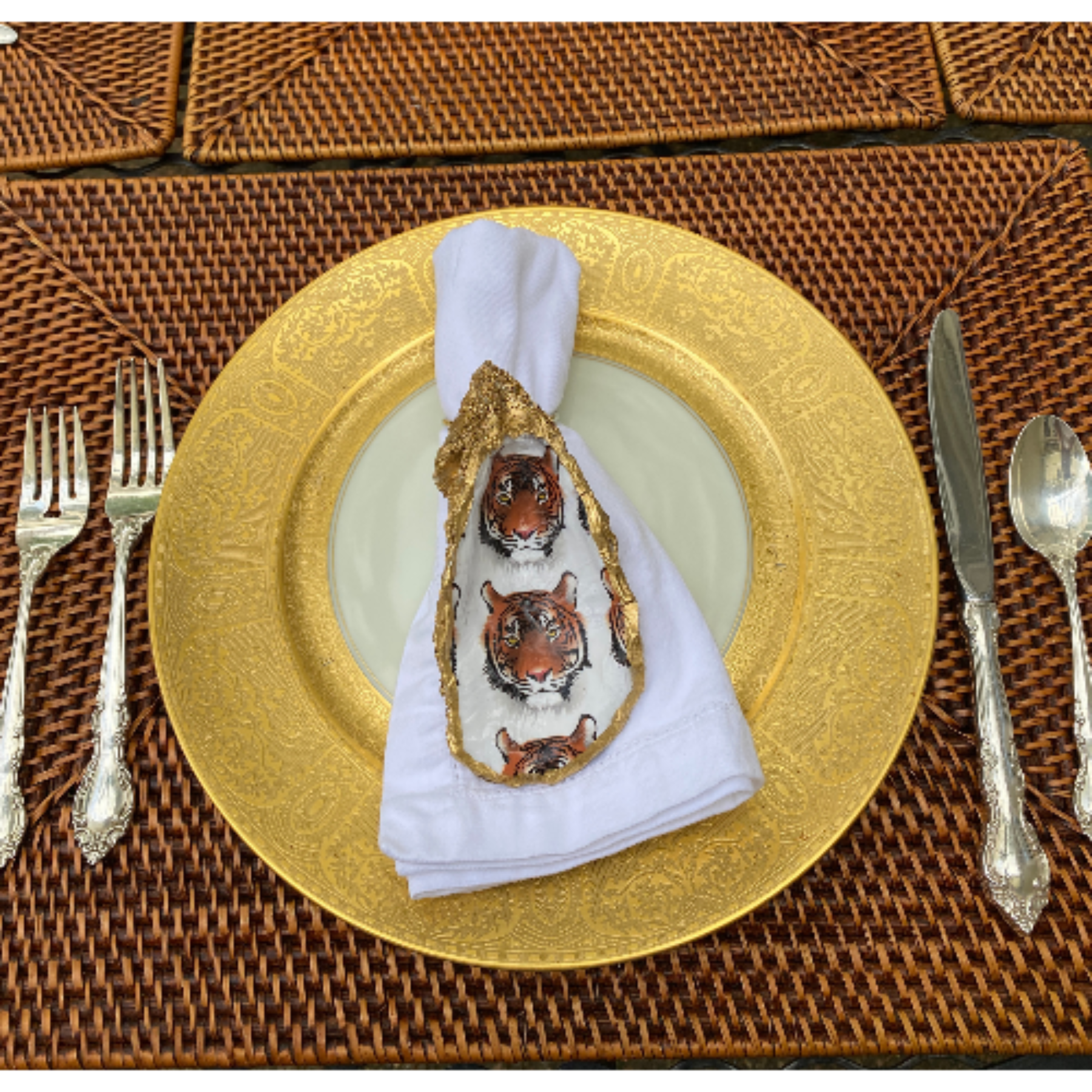 Gold leafed tiger head napkin ring on a white napkin, displayed on a golden plate with silverware on a wicker table.