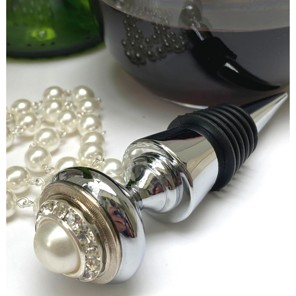 A silver wine bottle stopper with a large faux pearl set at the base, accompanied by clear crystals. The stopper is shown in a vertical position with a wine bottle in the background, and a string of pearls lying in front. Gift to celebrate 30th anniversary