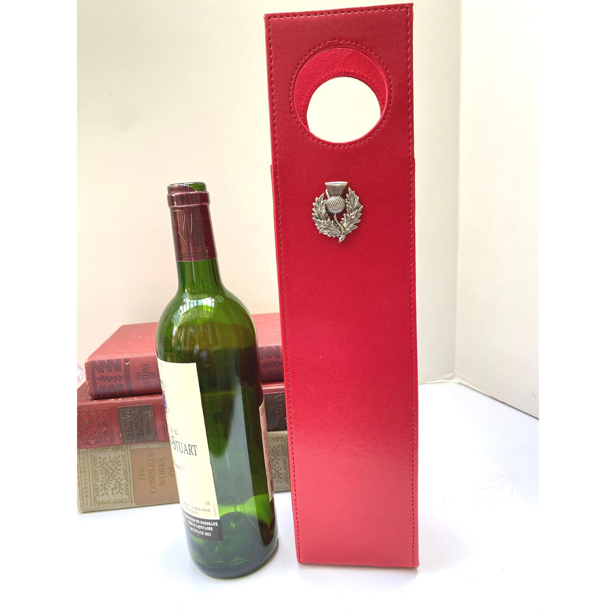 A red faux leather beverage carrier with a large silver thistle emblem, designed to hold a wine bottle, displayed in a collapsed state next to the bottle.